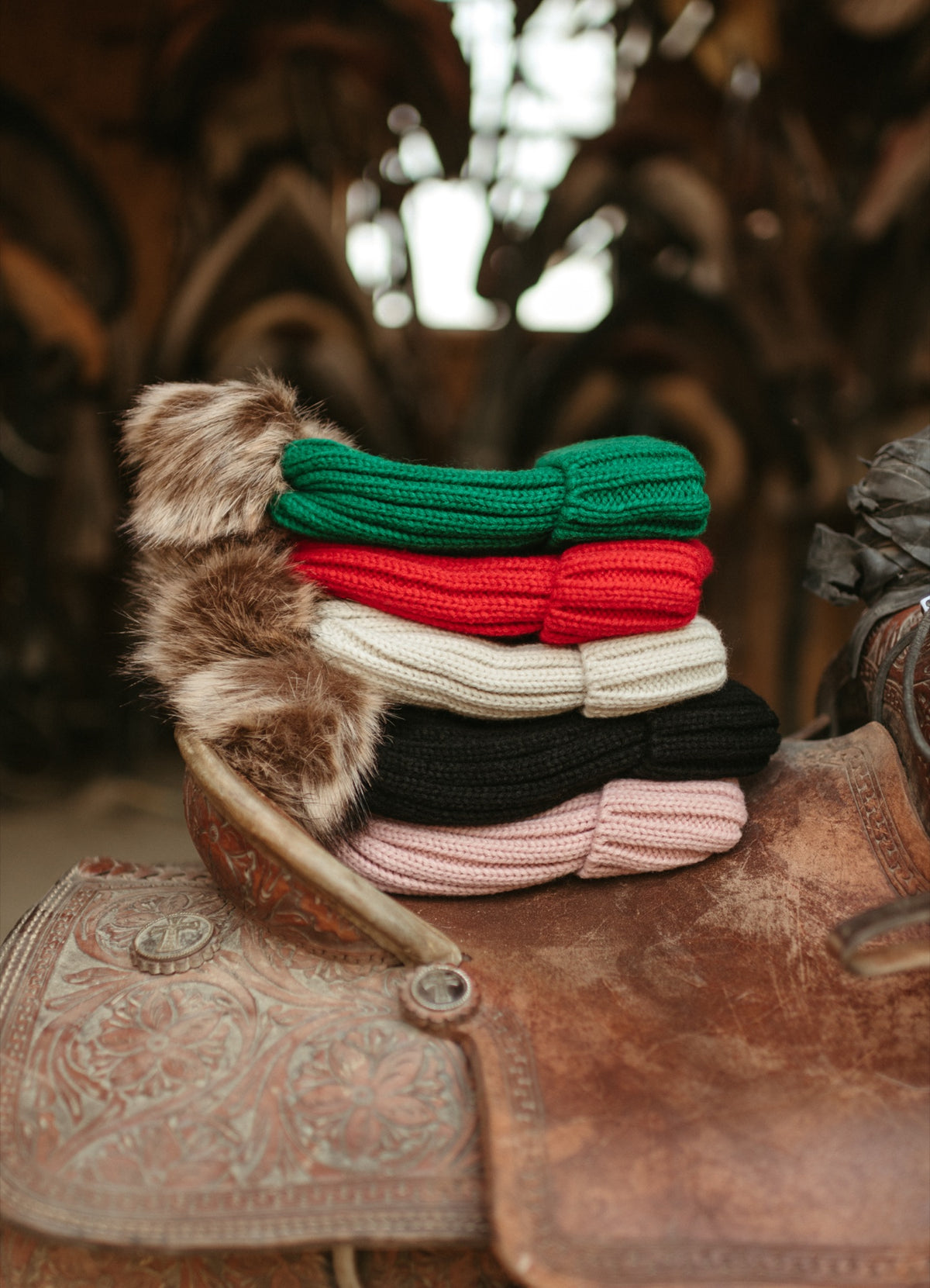 ASIAB knit beanies with faux-fur poms stacked on a worn leather saddle in the barn
